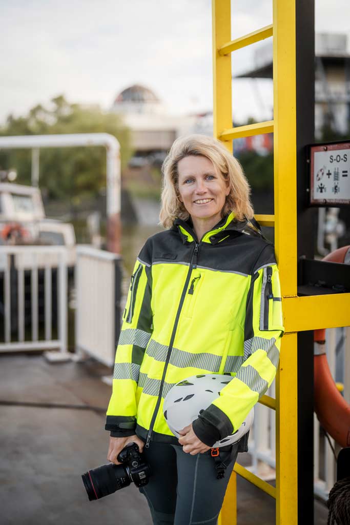 Britta Hilpert: Portraitbild mit PSA und Kamera: gelbe Warnjacke und Helm unter dem Arm an einer Leiter lehnend